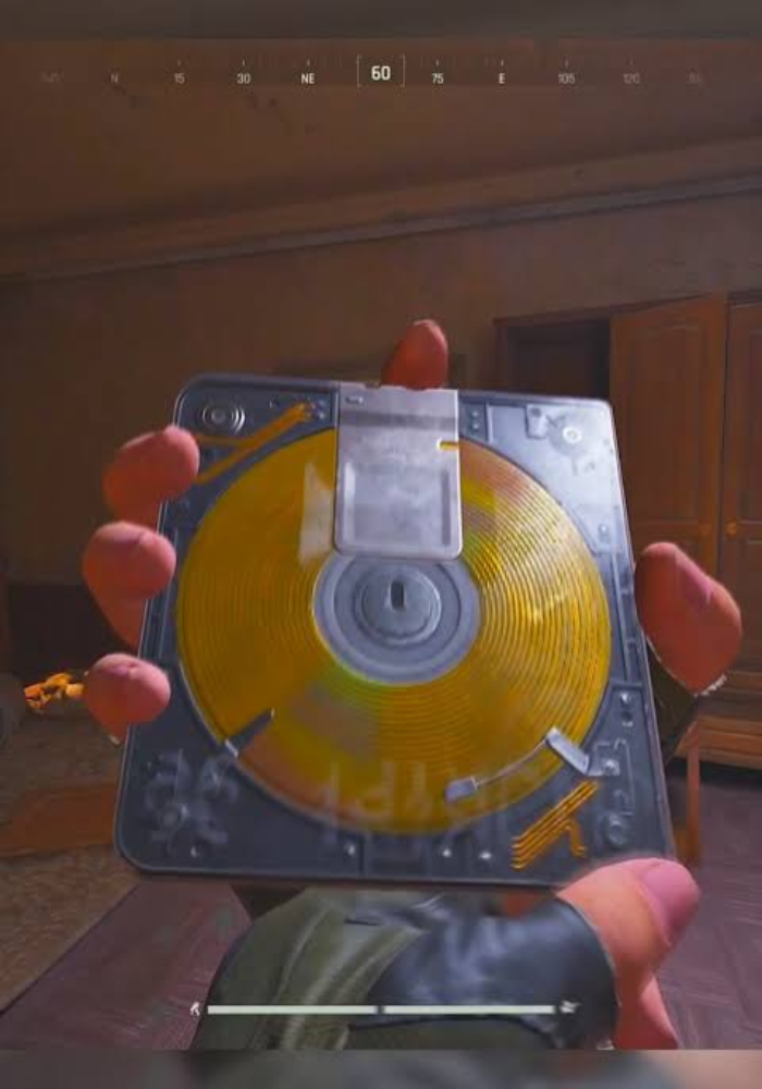 First-person video game screenshot of a hand holding a square case with a golden spiral disc inside, held up in a dimly lit room with wooden furniture. A compass HUD reading 60 NE runs across the top.