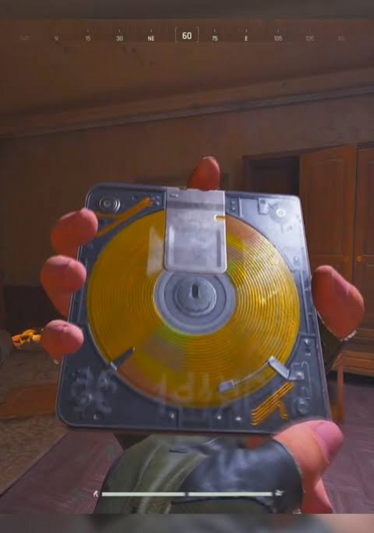 First-person video game screenshot of a hand holding a square case with a golden spiral disc inside, held up in a dimly lit room with wooden furniture. A compass HUD reading 60 NE runs across the top.